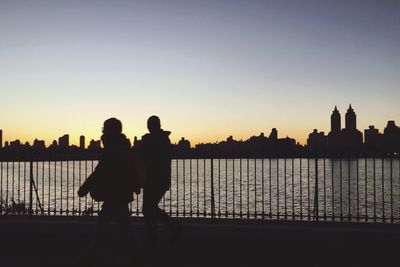 Silhouette people walking by river against cityscape during sunset