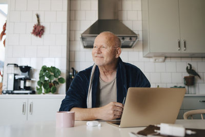 Smiling senior man with laptop looking away while sitting in kitchen at home