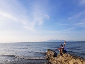 Man with arms raised sitting on rock looking at sea against sky