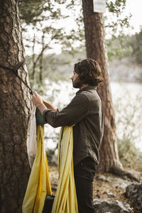 Side view of man hanging hammock on tree trunk in forest