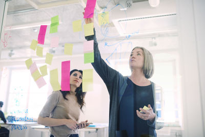 Female engineer discussing with colleague over adhesive notes on glass in creative office