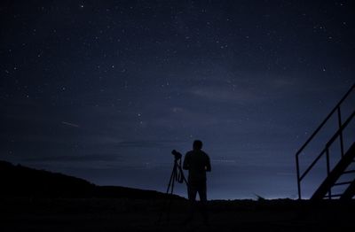 Silhouette man with camera and tripod standing against sky at night