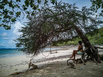 Young woman swinging in tree on beach against sky