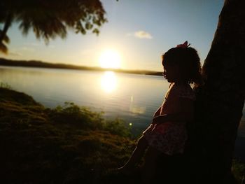 Side view of girl leaning on tree trunk by lake during sunset