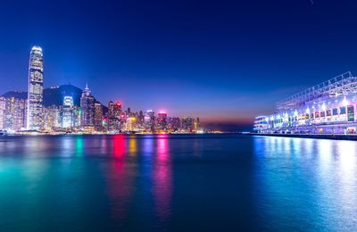 Illuminated buildings by river against blue sky at night