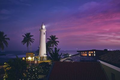 Illuminated building by lighthouse against sky at sunset