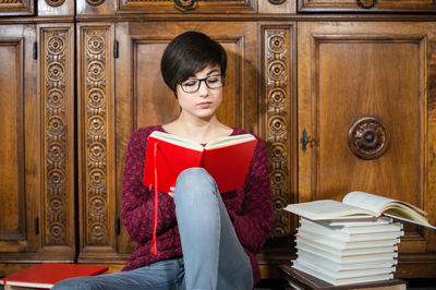 Young woman reading book while sitting against wooden furniture in library