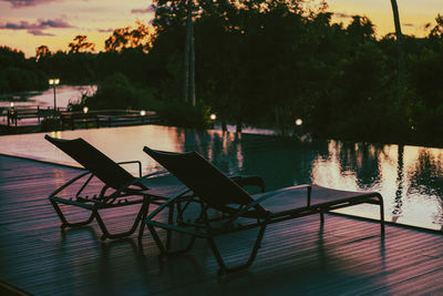 Empty chairs and tables at swimming pool against sky at sunset