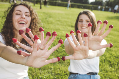 Playful female friends enjoying with fresh raspberries on fingers at park during sunny day