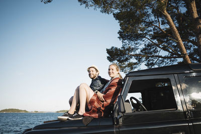 Couple sitting on jeep at lakeshore against clear blue sky