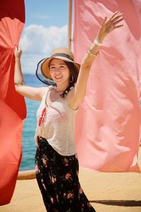 Pretty asian lady wearing a hat and accessories smiling waving at camera behind flags beach summer
