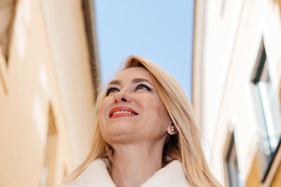 An upward angle captures a radiant older woman smiling against an urban sky