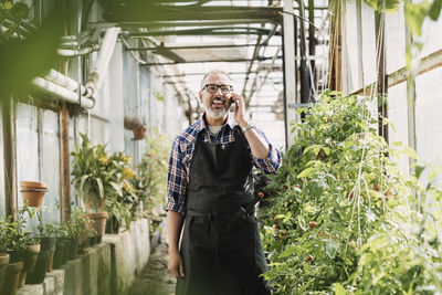 Happy gardener talking on mobile phone while standing in greenhouse