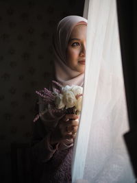 Close-up of young woman holding bouquet while standing by curtain at home