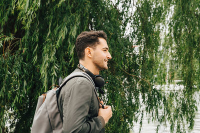 Side view of young man with backpack standing by tree in city