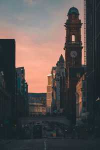 Traffic on road by buildings against sky during sunset