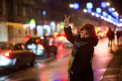 Portrait of woman showing peace sign while standing on street in city at night