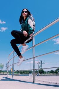 Low angle view of woman sitting on railing against blue sky