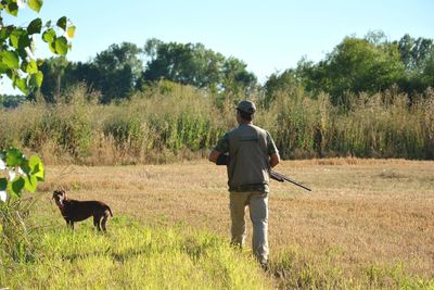 Rear view of man holding gun by dog on farm