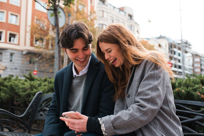 Positive young couple in love sitting on bench and discussing content on mobile phone while enjoying time together in city