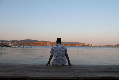 Rear view of man looking at sea while sitting on pier against clear sky