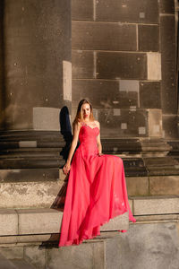 Full length of young woman in pink evening gown sitting against columns