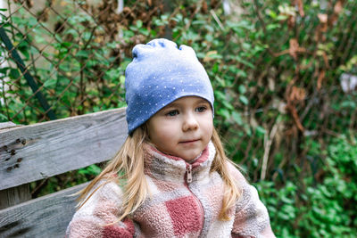 A cute preschool girl with long blond hair and blue eyes sitting on a wooden bench in the garden. 