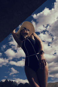 A portrait of a woman in a swimsuit, hat and sunglasses in summer on the riverbank against the sky