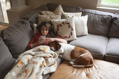 Tween girl looking at her phone while cuddling with hound dog on couch