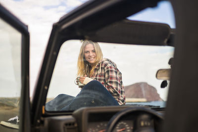 Portrait of young woman holding coffee cup while sitting on car hood