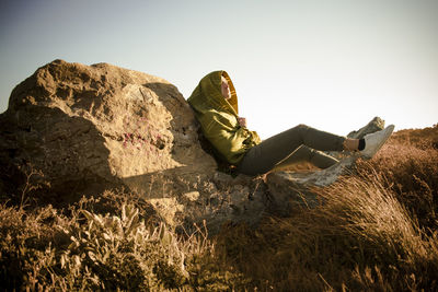 Side view of man with blanket sitting on rock against clear sky