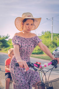 Young woman riding bicycle with boy sitting in background
