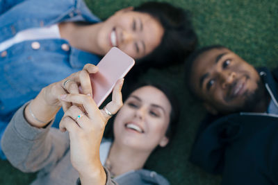 High angle view of smiling young woman taking selfie with friends while lying down on grass