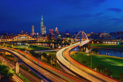 Light trails on road amidst illuminated buildings against sky at night