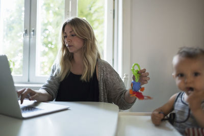 Mid adult mother using laptop while taking care of baby at dining table