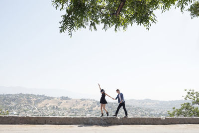 Excited young couple walking on retaining wall against clear sky