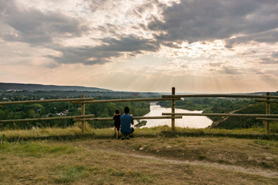 Rear view of father and son looking at river against sky from hill during sunset