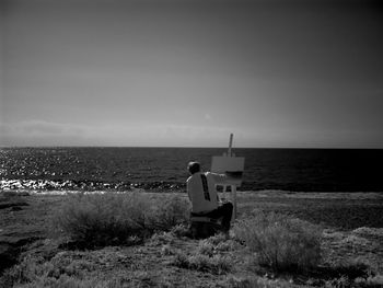 Rear view of man painting on canvas while sitting on field by sea against sky