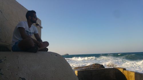 Man sitting on rock by sea against clear sky