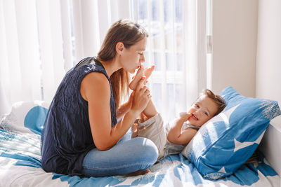 Young caucasian mother kissing boy toddler son feet heels legs. mom playing with child baby on bed 