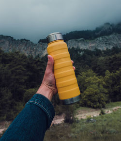 Male hand holding a steel yellow thermo bottle on nature background.