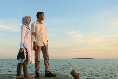 Couple standing on pier over sea against sky during sunset