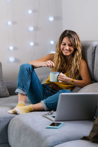 Smiling woman holding coffee cup while looking at laptop on sofa