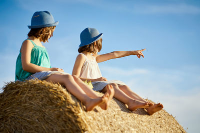 Low angle view of friends wearing hats sitting on hay bale against sky