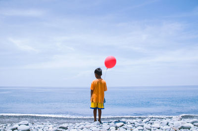 Rear view of boy with red helium balloon standing on shore at beach against sky