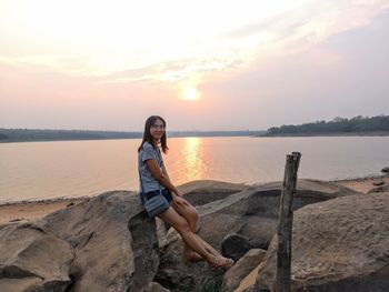 Portrait of young woman standing on rocks at beach against sky during sunset