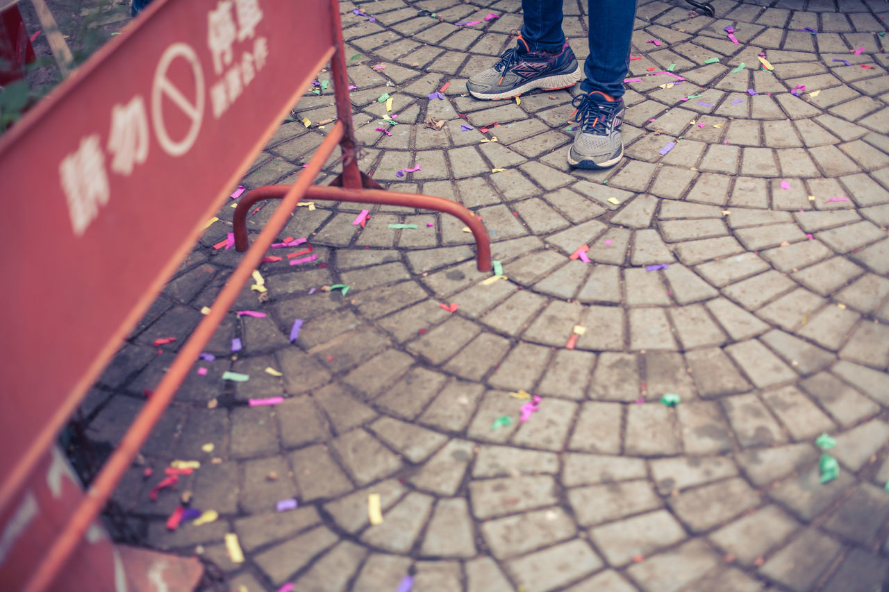 low section, real people, body part, human leg, footpath, human body part, high angle view, street, day, lifestyles, one person, city, outdoors, text, walking, shoe, standing, incidental people, men, paving stone, human foot