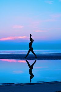 Silhouette mid adult woman exercising at beach against sky during sunset