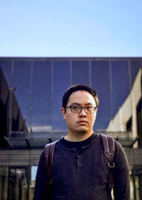 Portrait of young man standing against reflective glass facade and blue sky.