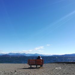 Rear view of woman sitting on bench by lake against blue sky during sunny day